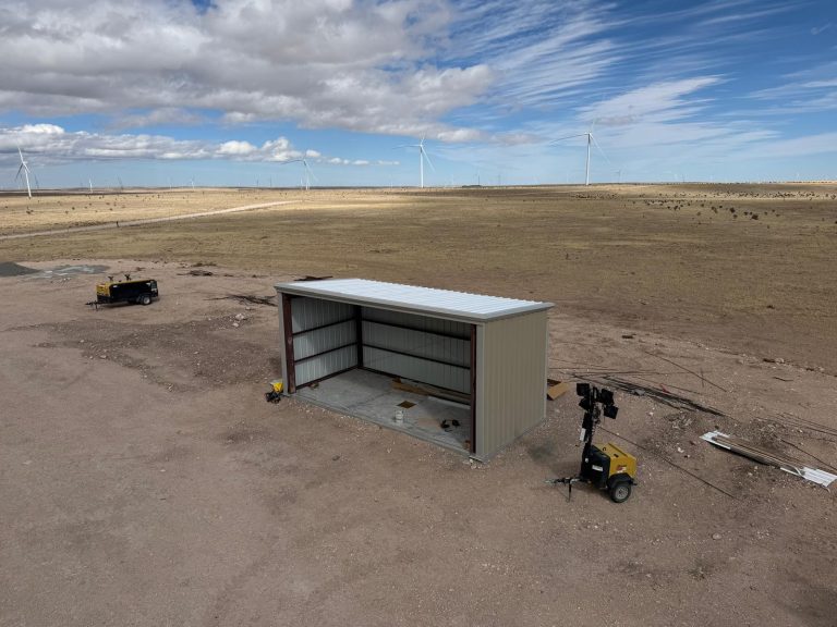 Three-sided steel shed with a light tower and compressor on prairie, with wind turbines on the horizon