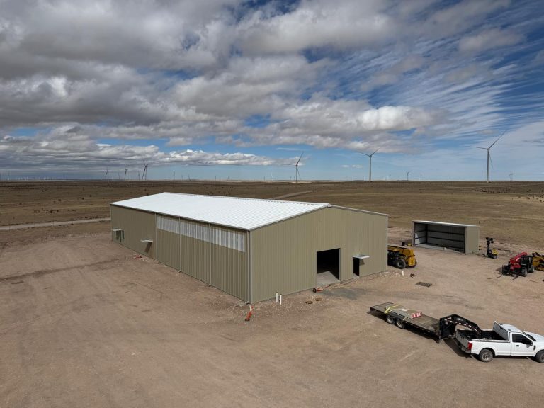 Aerial view of a tan steel building and an open-front shed, with a flatbed trailer, pickup and lifts