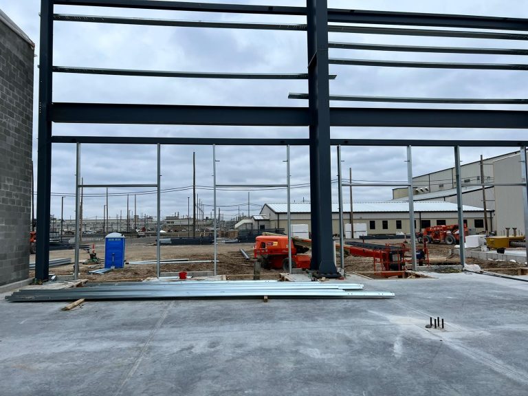 Dark steel frame and girts going up beside a concrete block wall, with lifts and buildings behind