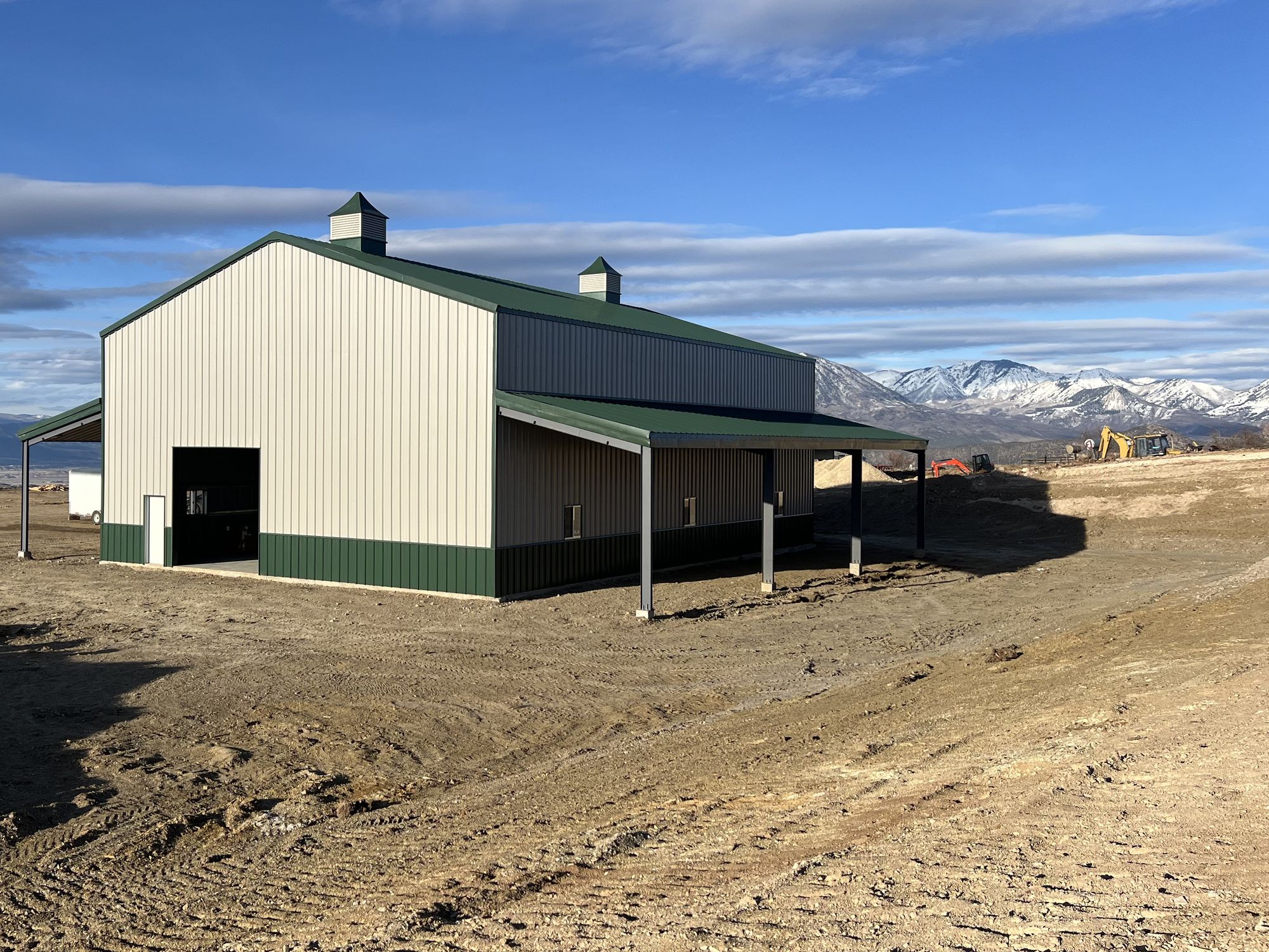 Cream steel barn with a green roof, green wainscot, cupolas along the ridge and lean-to sheds, with snowy mountains behind, near Hotchkiss, Colorado