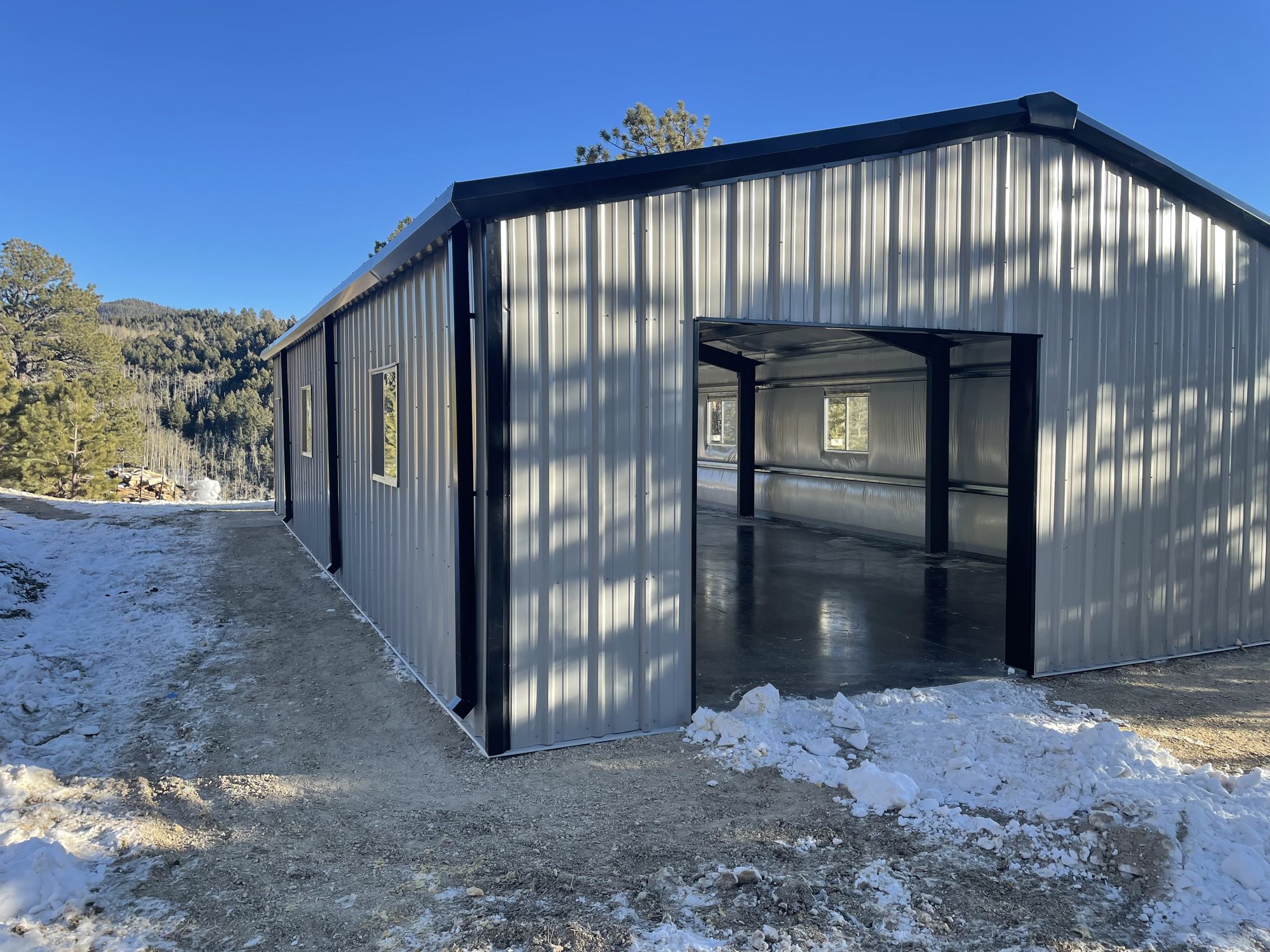 Gray steel shop with dark trim and a wide open door opening among pines, with snow on the ground, near Westcliffe, Colorado