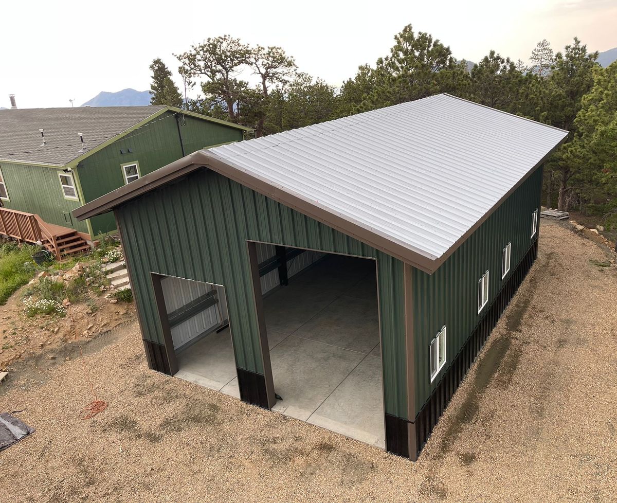 Green steel shop with two overhead door openings of different sizes and a silver roof, with mountains behind, near Golden, Colorado