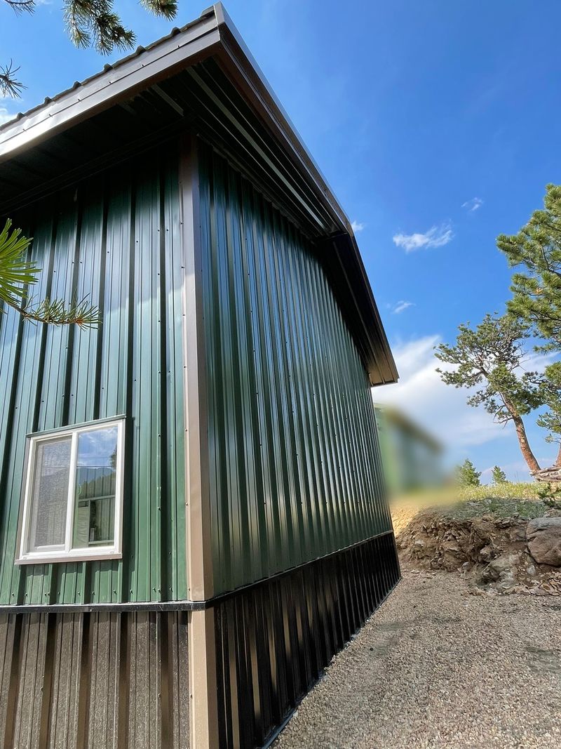 Corner of a green steel shop with tan corner trim, a dark wainscot, a white sliding window and a wide roof overhang, near Golden, Colorado