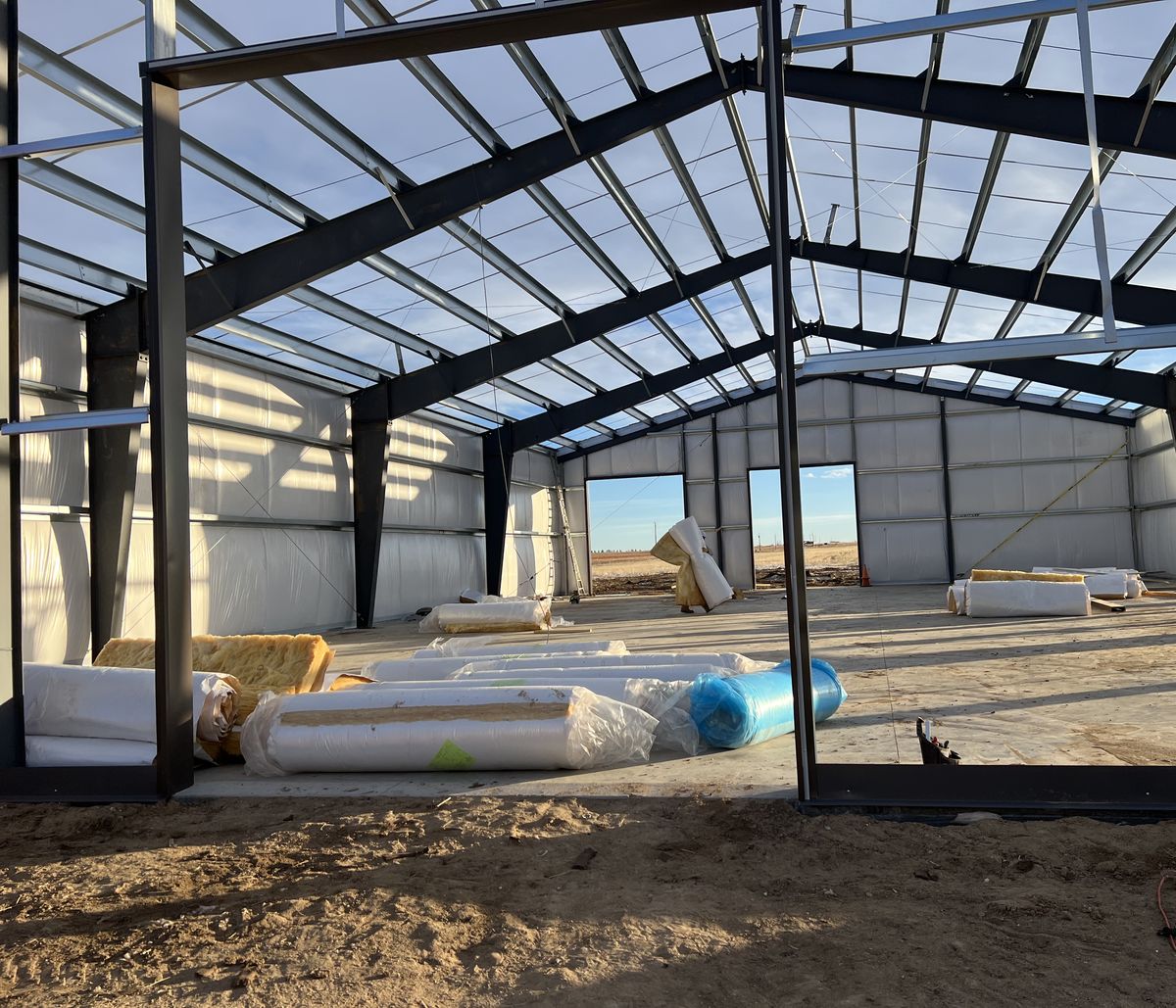 Inside a steel shop during erection: dark frames and galvanized purlins open to the sky, white-lined walls and rolls of insulation on the slab, near Byers, Colorado