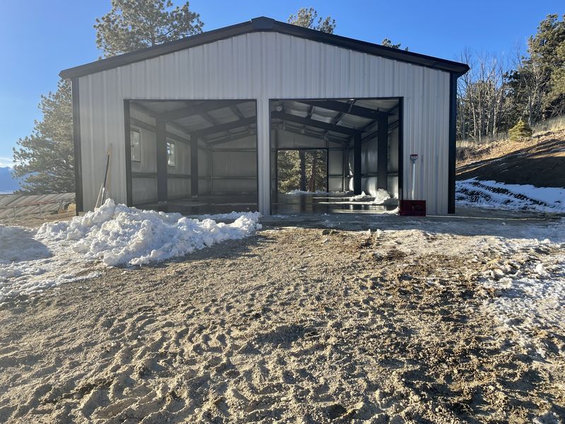 Silver steel shop with black trim and two large open bays showing dark frames and white insulation, with a snow pile outside, near Westcliffe, Colorado