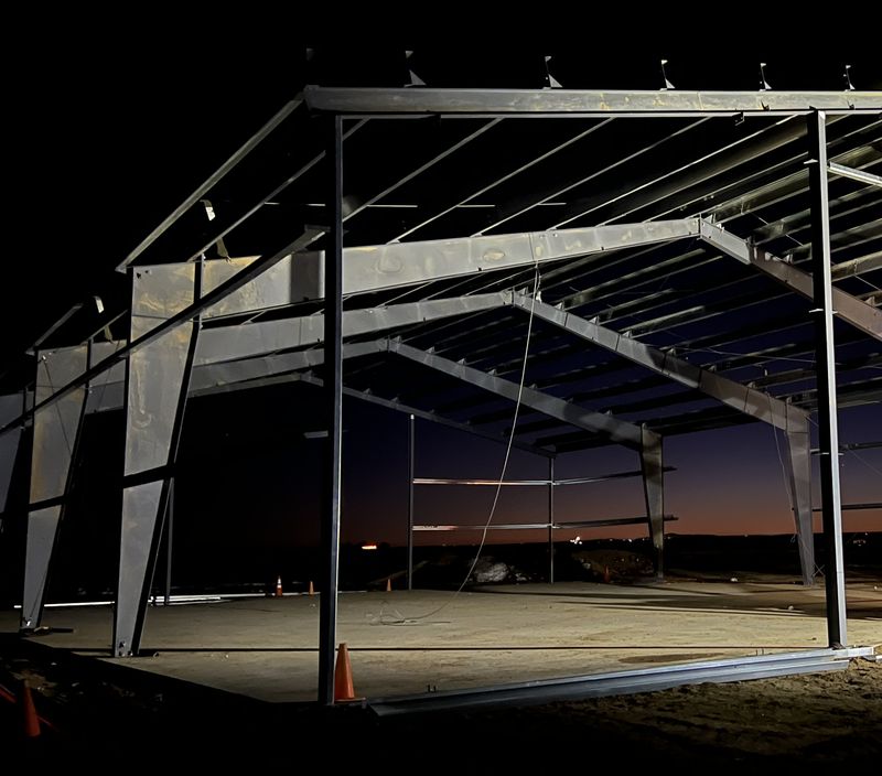 Steel shop frames and roof purlins standing at dusk under work lights, on a concrete slab with traffic cones, near Byers, Colorado