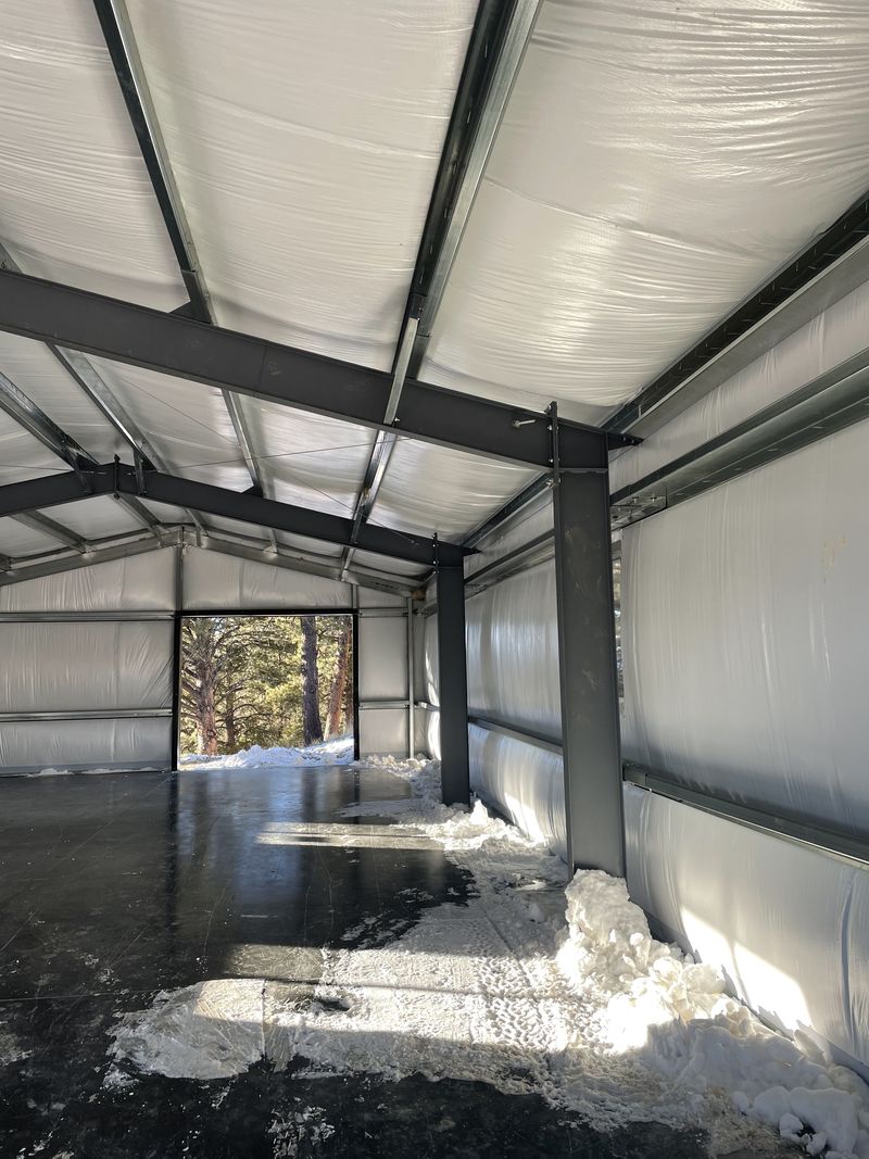 Inside a steel shop: dark frames, galvanized purlins and girts and white-faced insulation, looking toward an open end-wall door, near Westcliffe, Colorado