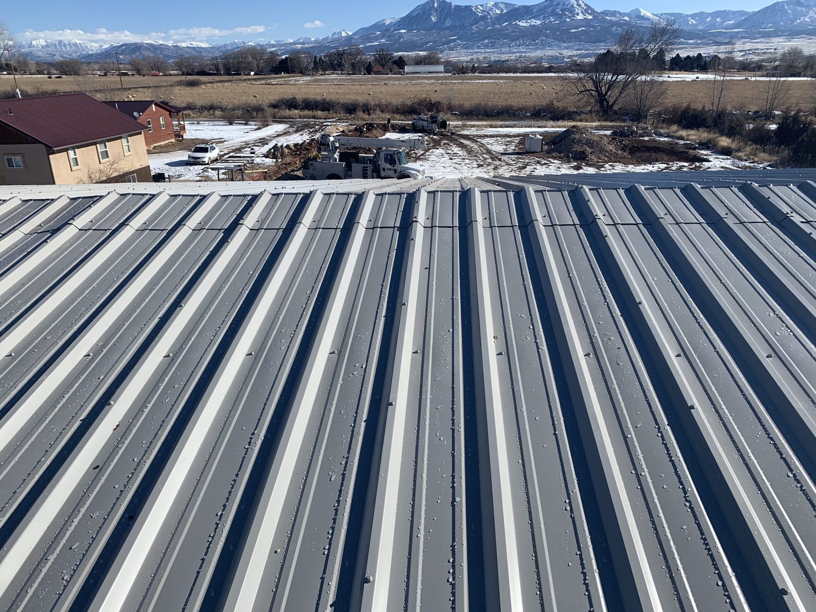 Gray metal roof panels on a steel building, seen from the roof, with snow-capped mountains behind