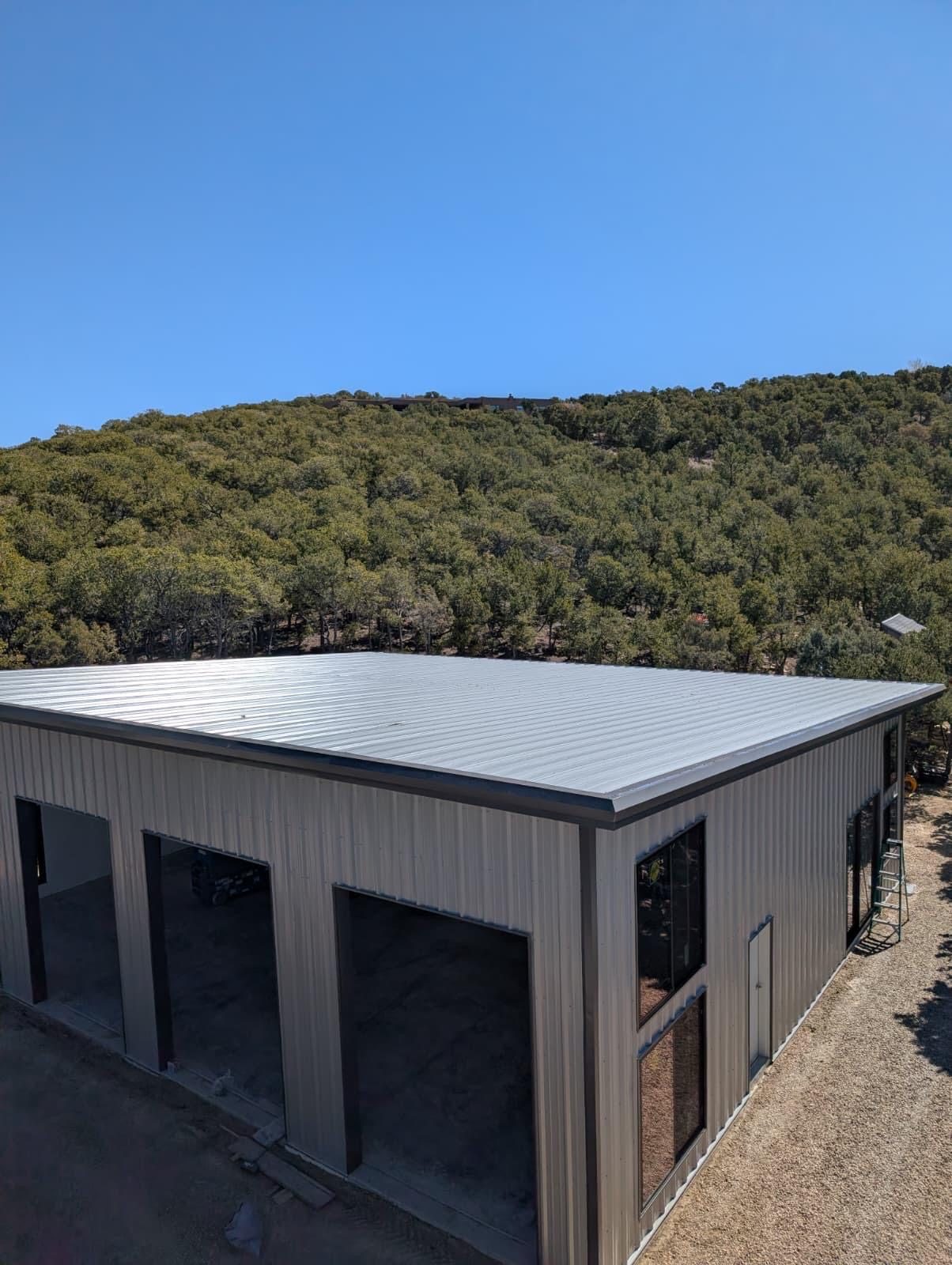 Light gray steel shop with three door openings and tall windows, below a tree-covered hillside
